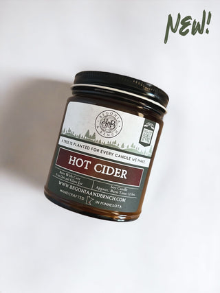 The Hot Cider™ - 9oz. Amber Jar Candle by Begonia & Bench® is presented in a brown glass jar and contains a soy fall candle. The label highlights the scent as featuring apple, pear, vanilla bean, and honey. It also includes the message: "A tree is planted for every candle we make," along with the website "www.begoniaandbench.com." The word "NEW!" is prominently displayed at the top right in green.