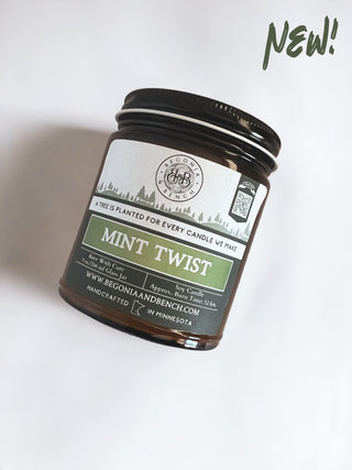 A 9oz. amber jar candle named "Mint Twist™" from Begonia & Bench® is showcased against a plain background. The label mentions that a tree is planted for every candle made. This handcrafted scented candle originates from Minnesota, with the word "NEW!" prominently displayed in green at the top right corner.