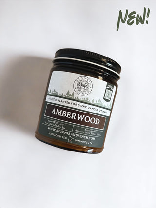 The 9oz Amberwood jar candle by Begonia & Bench® comes in a brown glass container with a black lid. The label, which reads "Amber Wood," mentions that a tree is planted for every candle made and features a QR code. Also displayed are the brand name, "Begonia & Bench®," and the website, www.begoniaandbench.com. The word "NEW!" is highlighted in green at the top right corner.