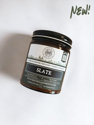 A brown amber glass jar candle with a black lid labeled "Slate™ - 9oz. Amber Jar Candle" by Begonia & Bench®. The label mentions, "A Tree Is Planted For Every Candle We Make." Approximate burn time: 45 hours. The words "NEW!" are written in green in the top right corner. This handcrafted fall-scented candle is made in Minnesota.