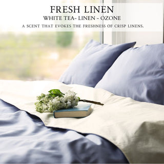 A neatly made bed adorned with light blue and white linens hosts a book and a white flower bouquet. Sunlight pours in from a nearby window, illuminating the scene. The ambiance is enhanced by the Fresh Linen™ - 12oz Finely Crafted Scented Candle By Begonia & Bench®, placed beside the bouquet, emanating a fresh linen aroma. The text at the top reads, "FRESH LINEN WHITE TEA - LINEN - OZONE" and "A SCENT.