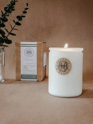 A lit white candle in a frosted glass container, labeled "Fresh Linen™ - 12oz Finely Crafted Scented Candle By Begonia & Bench®," sits on a brown surface. Behind it to the left, a brown cardboard box describes the handcrafted premium botanical wax scented candle. A sprig of greenery in a slim glass vase adds a touch of nature's elegance.