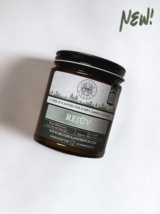 A 9oz amber glass candle jar with a black lid labeled "Rejüv™" from Begonia & Bench®. This scented candle mentions "A tree is planted for every candle we make" and "Handcrafted in Minnesota." The words "NEW!" are written in green in the top right corner. The fall candle is positioned against a light background.