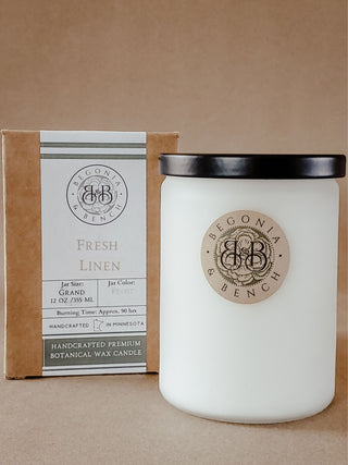 A Fresh Linen™ - 12oz Finely Crafted Scented Candle by Begonia & Bench® in a white, frosted glass jar with a black lid sits in front of a brown box. The jar features a circular logo with "Begonia & Bench," while the box displays the matching logo along with text that reads "Fresh Linen," 12 oz / 355 ml, approximate burn time of 80 hours, and handcrafted in Minnesota.