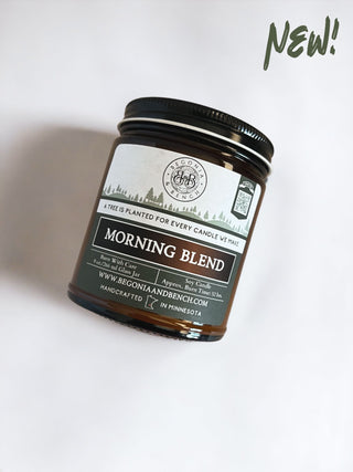 A dark amber glass jar of Morning Blend™ - 9oz. Amber Jar Candle from Begonia & Bench® with a black lid. The label features a green tree line design and states "A tree is planted for every candle we make." Infused with aromas of Arabica coffee and vanilla, the candle is handcrafted in Minnesota. Text reads "NEW!" in green in the top right corner.