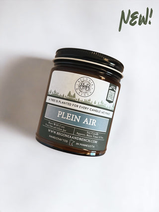The Plein Air™ 9oz. Amber Jar Candle by Begonia & Bench® comes in a brown glass jar. The label reads: "A tree is planted for every candle we make," and includes the website www.begoniaandbench.com. It also mentions that the product is handcrafted in Minnesota, with "NEW!" highlighted in green at the top right corner.
