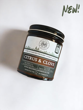 An amber glass jar candle labeled "Citrus & Clove - 9oz." from Begonia & Bench® sits on a light background. The label features trees and includes the message, "A tree is planted for every candle we make." It also highlights the burn time (35+ hours) and lists the website www.begoniaandbench.com. Text at the top right reads "NEW!