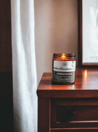 A scented candle labeled "Amberwood - 9oz. Amber Jar Candle - By Begonia & Bench®" in a brown glass jar sits on a wooden nightstand next to a white curtain. The background includes a framed picture partially visible to the right, creating a warm and cozy ambiance.