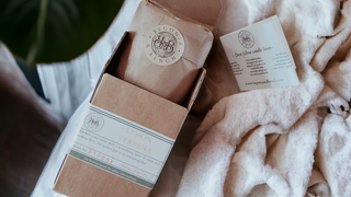 A brown cardboard box labeled "Lemon Verbena" from Begonia & Bench is partially opened, revealing a brown paper package inside. Next to the box is an unfolded card with text and the brand's logo. The items are placed on a soft, light-colored fabric.