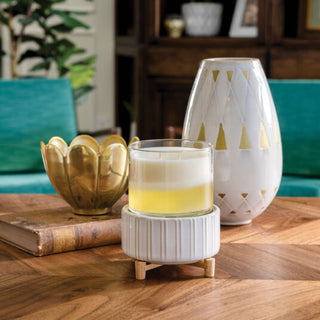 A cozy setting features a Classic 2-in-1 Candle Warmer, gently illuminating the space while releasing soothing scents from wax melts. Nearby, a large white vase with gold triangle accents and a brass lotus-shaped bowl adorn the wooden table. Vintage books and teal chairs enhance the warm, inviting atmosphere.