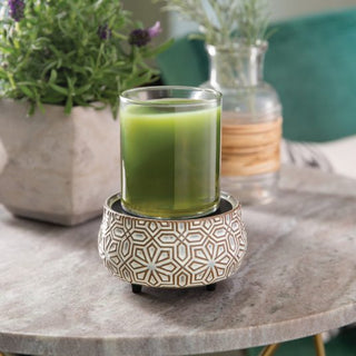 A green jar candle in a glass holder sits on the Classic 2-in-1 Candle Warmer, showcasing its elegant geometric design. The warmer is placed on a round marble table, while blurred potted plants—one leafy and another lavender—enhance the cozy, inviting ambiance in the background.