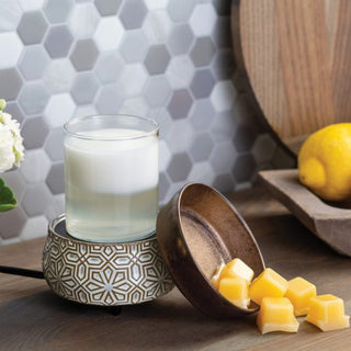A Classic 2-in-1 Candle Warmer with a patterned base holds a jar candle filled with white wax. Close by, a brown bowl is filled with aromatic wax melts. In the backdrop, a hexagonal tile backsplash enhances the scene of a wooden tray that displays a lemon and white flowers, all situated on a wooden surface.