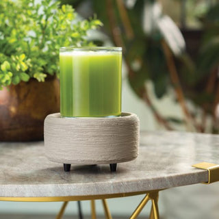 A Classic 2-in-1 Candle Warmer in a glass holder is displayed on a textured beige base with small legs atop a marble tabletop. The scene is enhanced by blurred green plants in the background, creating a calming and natural ambiance, while the gentle scent of fragrance tarts adds an elegant touch alongside gold accents.