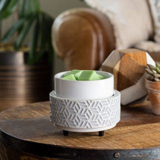 A white, textured Classic 2-in-1 Candle Warmer featuring a geometric pattern rests on a wooden table, holding aromatic wax melts. Nearby, there's a small potted succulent and a decorative wooden piece. A brown leather sofa and houseplant are softly blurred in the background, enhancing the cozy atmosphere.