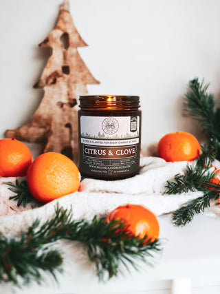 A lit Citrus & Clove - 9oz. Amber Jar Candle by Begonia & Bench® is placed on a white cloth surrounded by tangerines and pine branches. The background features a rustic, wooden Christmas tree decoration, evoking a cozy, winter holiday ambience.