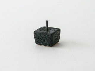 A small, textured black cube with a rough, porous surface sits on a plain white background. A slender, vertical black rod, reminiscent of traditional Japanese candle designs, is inserted into the top center of the cube, extending upwards. The rod appears to be made of the same material as the cube. This elegant piece is part of the DAIYO Rice Wax Candle Gift Box.