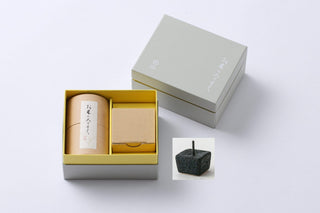 A neatly opened gray gift box reveals a cylindrical container with Japanese text and a small brown box inside. The lid of the box shows minimalist Japanese writing and a logo. Beside the box, there is a black, square incense holder with a stick incense placed upright, enhancing the ambiance for your DAIYO Rice Wax Candle Gift Box experience.