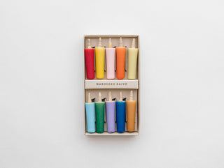 A box of ten colorful Japanese candles arranged in two rows of five. The top row includes red, light green, yellow, pink, and orange candles. The bottom row features blue, purple, green, light blue, and dark yellow candles. Made from natural wax with a washi paper label reading "DAIYO Vivid Color Rice Wax Candles." The background is white.