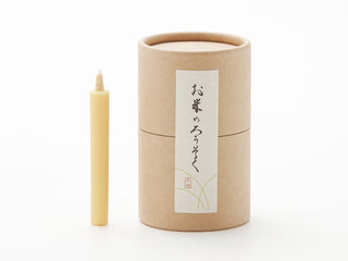 A small, unlit, yellow DAIYO Rice Wax Candle stands upright next to a cylindrical, brown cardboard box. The box has a Japanese label featuring black and red kanji characters on a white background with some decorative lines. This pure vegetable-based Japanese candle appears to be packaged inside the box.
