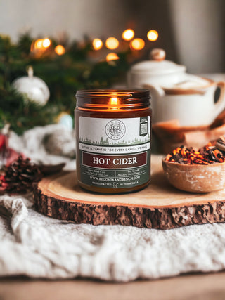 A glowing Hot Cider™ 9oz. Amber Jar Candle by Begonia & Bench® is situated on a rustic wooden slice. The background showcases a blurred Christmas tree adorned with lights, pinecones, a teapot, and a wooden bowl brimming with spices on a light-colored cloth, creating a cozy holiday ambiance.