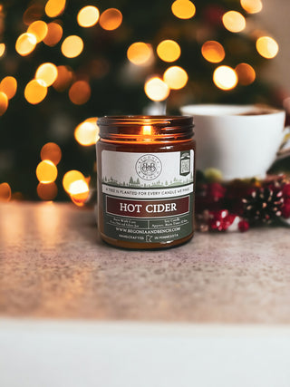 A lit Hot Cider™ candle from Begonia & Bench® in a 9oz. amber jar sits on a marble surface. The background is adorned with warm, blurred bokeh lights, possibly from a Christmas tree. A white coffee cup and festive holiday decor, including red berries and greenery, are also visible in the background.