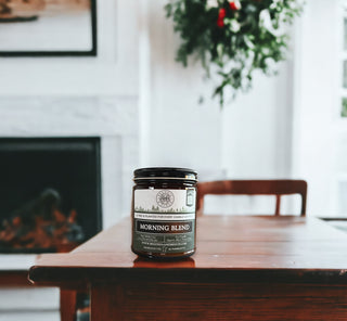 An amber jar labeled "Morning Blend™ - 9oz. Amber Jar Candle - By Begonia & Bench®" is placed on a wooden table. The blurred background reveals a white fireplace, some greenery, and part of a chair. The label on the jar features text and a logo, indicating it may be related to coffee or possibly an autumn candle. Light filters through a nearby window.