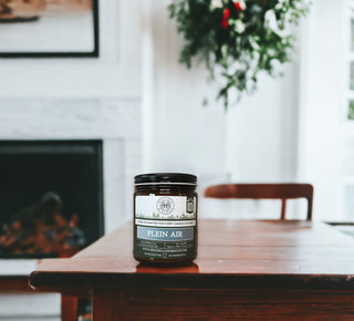 A 9oz. amber jar candle labeled "Plein Air™ - By Begonia & Bench®" sits centered on a wooden table. The background shows a fireplace with low flames and a white mantle adorned with greenery. To the right, part of a chair and blurred green foliage are visible, creating a cozy indoor atmosphere.