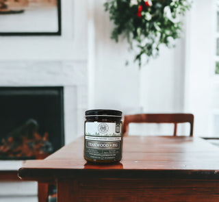 A 9oz amber jar candle labeled "Teakwood & Fig™" by Begonia & Bench® sits prominently on a wooden table, with a blurred background featuring a fireplace and greenery. This fall candle is crafted with soothing scents perfect for relaxation and unwinding.
