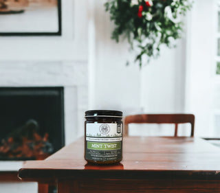 A 9oz. amber jar candle labeled "Mint Twist™ - By Begonia & Bench®" is centered on a wooden table. The room is bright with a white fireplace and a green plant hanging in the background. The candle, which has a green label with white text and a logo, contributes to the cozy and inviting atmosphere.