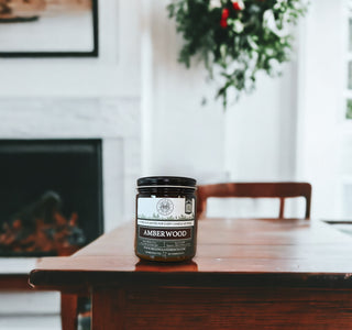 A 9oz. Amber Jar Candle by Begonia & Bench®, featuring the Amberwood scent, sits on a polished wooden table indoors. The black candle jar showcases a label adorned with white and gold text. In the background, there's a blurred view of a white fireplace and some greenery, adding to a cozy and warm ambiance.