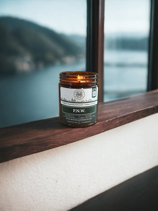 A 9oz. amber jar candle labeled "P.N.W." by Begonia & Bench® with a green and white design sits on a wooden windowsill. The window offers a view of a misty, forested mountain and a large body of water, creating a serene and cozy atmosphere.