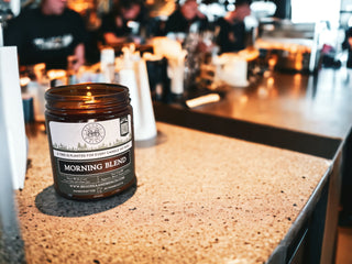 A brown 9oz. amber jar candle labeled "Morning Blend™ - By Begonia & Bench®" sits on a speckled granite counter in a bustling cafe. In the background, a barista prepares drinks while several patrons are seated along the counter, contributing to a warm and busy atmosphere. The candle's label also mentions that a tree is planted for each candle sold.