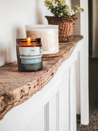 A Plein Air™ 9oz. Amber Jar Candle by Begonia & Bench® sits on a rustic wooden shelf. Behind it, a white textured container with small green plants in a woven basket rests on another shelf in the background, creating a cozy and natural ambiance in the room.