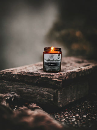 A small brown jar candle labeled "Slate™ - 9oz. Amber Jar Candle - By Begonia & Bench®" is lit, placed on a weathered, rough wooden surface. The scented candle emits a warm, cozy glow, standing out against the dark, blurred background and creating an atmosphere of warmth and tranquility.