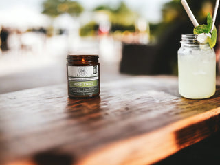 A small jar labeled "Mint Twist™ - 9oz. Amber Jar Candle - By Begonia & Bench®" sits on a wooden table outdoors. The jar, a scented candle, exudes freshness. Next to it is a mason jar with a light green beverage, garnished with a sprig of mint and a bamboo straw. The background is blurred but shows a sunlit outdoor setting.