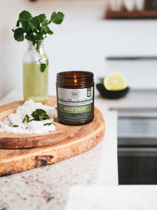 A scented candle in a 9oz. amber jar labeled "Mint Twist™" by Begonia & Bench® sits on a wooden tray filled with sea salt, with a sprig of mint beside it. In the background, a bottle of mint-infused liquid and a bowl containing a halved lime are placed on a kitchen countertop. The setting is bright and minimalistic.