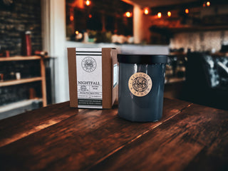 A fall candle and its packaging sit on a wooden table. The 12oz finely crafted scented candle by Begonia & Bench®, labeled "Nightfall™," boasts a black lid and container. Beside it rests a brown and white box, also named "Nightfall™," detailing fragrance notes of blackcurrant and birchwood. The background showcases a warmly lit, cozy room.
