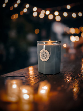 A Nightfall™ - 12oz Finely Crafted Scented Candle By Begonia & Bench® in a glass jar with a printed label is lit, emitting a warm glow in a dimly lit setting. The candle sits on a wooden surface with a few more candles in the foreground, out of focus. In the background, various out-of-focus string lights create a cozy atmosphere.