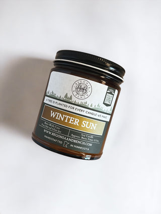 A handcrafted scented candle from Begonia and Bench®, the "Winter Sun™ - 9oz. Amber Jar Candle," features a white label with green and orange accents, indicating it is made in Minnesota. Additionally, for each candle sold, a tree is planted. The brown glass jar has a black top, standing out against a crisp white background.