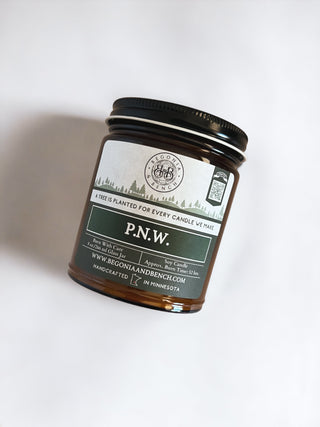 A photo of a 9oz amber glass jar candle with a black lid labeled "P.N.W." The label features a forest and mountain design and states "A tree is planted for every candle we make." This scented candle by Begonia & Bench® is handcrafted in Minnesota. Visit www.BEGONIAANDBENCH.COM.