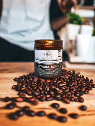 A lit brown fall candle labeled "Morning Blend™ - 9oz. Amber Jar Candle - By Begonia & Bench®" sits atop a pile of coffee beans on a wooden surface. The background is blurred, featuring a person wearing a light-colored shirt and green plants. Text on the candle notes that a tree is planted for every candle purchased.