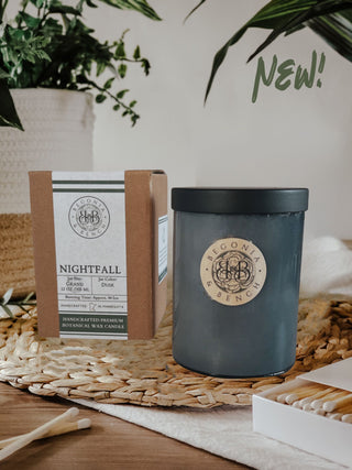 Image of a dark blue fall candle with a cork lid next to its box on a woven mat. The candle and box are labeled "Begonia & Bench" and "Nightfall™ - 12oz Finely Crafted Scented Candle By Begonia & Bench®," describing it as a handcrafted premium botanical wax scented candle with cedar and musk scents. The word "NEW!" is written in green in the top right corner.