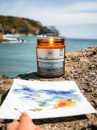 A lit "Plein Air™ - 9oz. Amber Jar Candle" by Begonia & Bench® rests on a rocky surface by the sea. A hand holds a piece of watercolor paper with a colorful painting, depicting blue sky and yellow-orange hues, in front of the scented candle. The background showcases a scenic coastline with cliffs and the ocean.