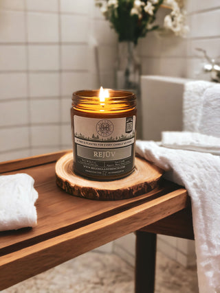 A lit Rejüv™ 9oz. amber jar candle by Begonia & Bench® sits on a round wooden coaster on a wooden surface in a bathroom. The background features white tiled walls, a blurred white bathtub, folded white towels, and a vase with white flowers.