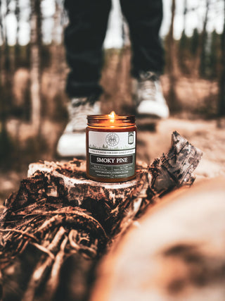 A lit Smoky Pine™ - 9oz. Amber Jar Candle by Begonia & Bench® is placed on a piece of tree bark in a forest setting. The background shows blurred trees and a person's lower legs and feet, wearing black pants and white shoes, standing on the tree bark.