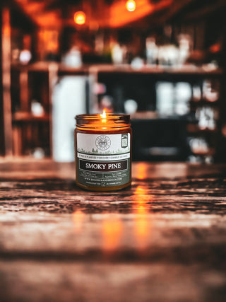 A lit Smoky Pine™ - 9oz. Amber Jar Candle by Begonia & Bench® sits on a rustic wooden surface. The background is a cozy, blurred interior with warm lighting and shelves stocked with various items, creating a warm and inviting atmosphere.
