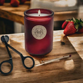 A lit red candle, labeled "Strawberry Shortcake - By Begonia & Bench®," sits in a frosted glass jar atop a wooden surface. Nearby are fresh strawberries, black wick trimmers, and a lit matchstick. This Strawberry Shortcake candle exudes warmth, crafting a cozy atmosphere with its inviting glow.