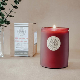 A lit red candle in a glass jar, adorned with the "Begonia & Bench" logo, rests on a surface, releasing the luxurious aroma of their Strawberry Shortcake creation. Adjacent to it, a charming box bearing the name "Strawberry Shortcake - By Begonia & Bench®" complements the scene. A small branch with green leaves enhances this picturesque setup against a beige background.