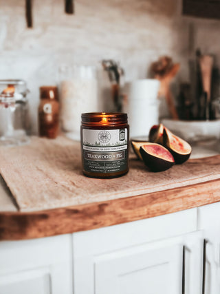 A lit Teakwood & Fig™ 9oz. Amber Jar Candle by Begonia & Bench® sits on a wooden surface in a cozy kitchen. Behind it on the countertop are various jars and containers. Two halved figs rest beside the fall candle, and brushes and utensils are visible in the softly blurred background.