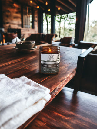 A 9oz. Amber Jar Candle labeled "Tobacco & Cedar" by Begonia & Bench® rests on a rustic wooden table next to a white cloth in a cozy, cabin-like setting. The background features dim lighting, wooden walls, and comfortable seating, creating a warm, inviting atmosphere. Trees are visible through the windows.